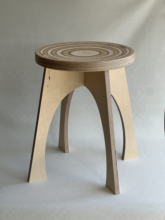 A birch plywood stool. Concentric rings machined into the round seat cut through the laminations, exposing the ply lines as circles, and the edge of the seat and the arched legs show their stacked plies.